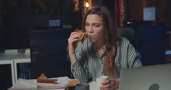 Frustrated Woman Office Manager Eatting Sandwich and Drinking Coffee at Night Office. Exhausted Girl