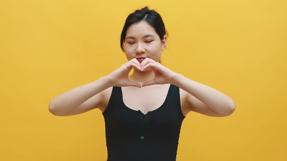 Happy Young Asian Woman Making Heart Shape with Her Hands. Isolated on Yellow Background