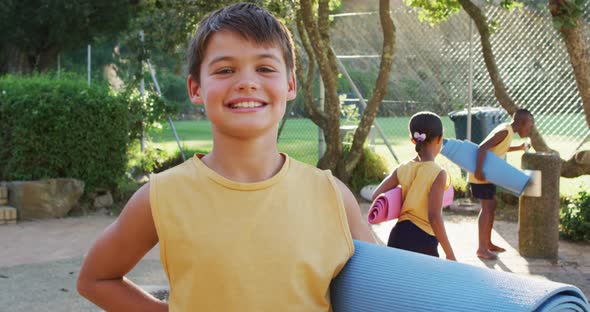 Portrait of happy caucasian schoolboy holding yoga mat after yoga lesson outdoors