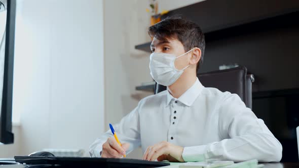 A Young Man in a Medical Mask Performs Homework at a Computer
