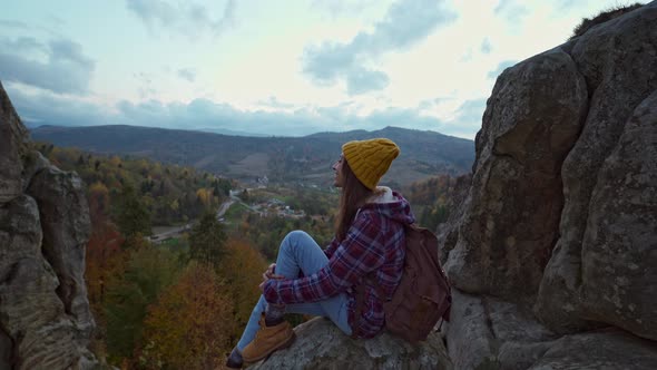 Girl Hiker in Checkered Jacket and Hat with Backpack Sits on Edge of Cliff and Enjoing Beautiful