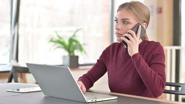 Young Woman with Laptop Talking on Smartphone in Office