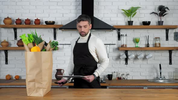 Man in black apron puts knife and board for cutting vegetables on kitchen table