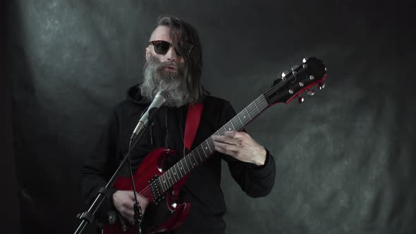 Bearded hairy rock musician in retro horn-rimmed sunglasses plays on red electric guitar