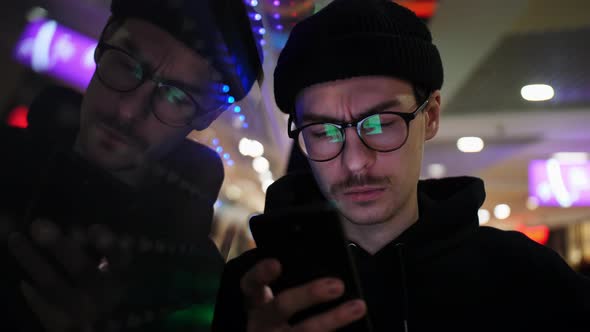 A Man with Glasses Looks at His Phone Near an Animated Light in a Shopping Mall