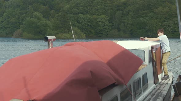 Young man aboard wooden boat wipes dust off wooden liveboard boat