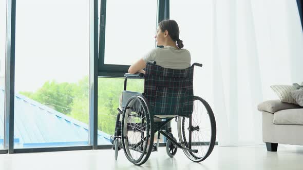 Smiling Young Woman in Wheelchair Relaxing at Home Near Window and Looks Outside