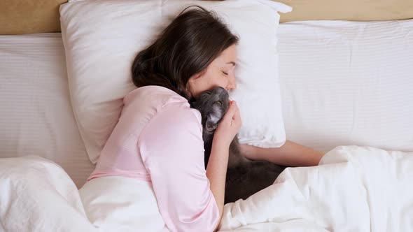 Relaxed Woman in Pale Pink Pajama Pets Sleepy Cat in Bed