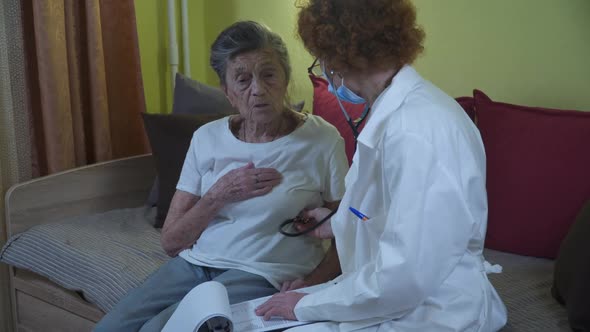 Mature Woman Doctor Holding Stethoscope Examining Senior Grandma Patient at Home