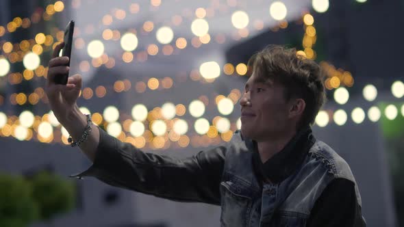 Young Man Holds a Smartphone in Her Hand and Takes a Selfie Outside in a Modern Residential Area