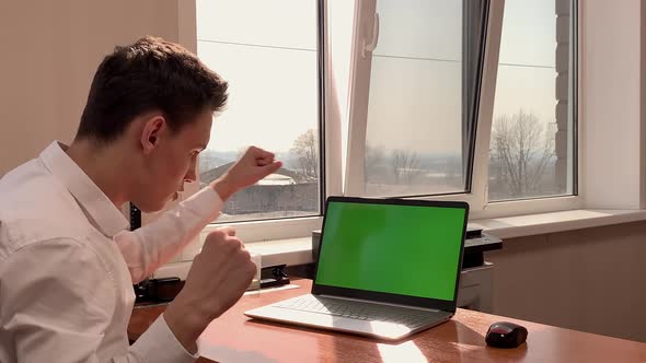 A Young Man Enjoys Success at Work Sitting at a Desk in Front of a Computer with a Green Screen