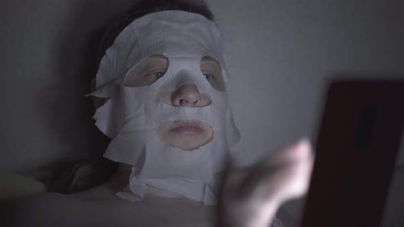 Young Woman in Regenerating Sheet Mask Lies in Dark Room