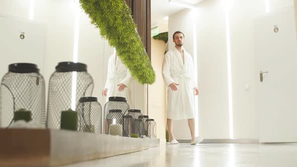 Young Man in White Robe Walks Down the Hall of Luxurious Spa Center Talking on Mobile Phone.