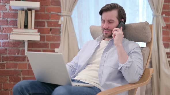 Casual Young Man with Laptop Talking on Smartphone on Sofa