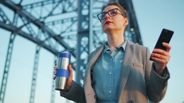 Woman in a Coat Walking Around the City in the Early Morning Drinking Coffee and Using Smartphone