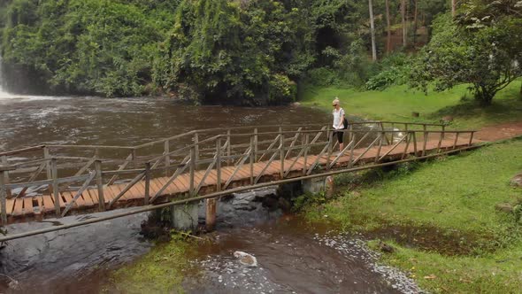 Aerial shot orbiting around a young blonde woman crossing an old wooden foot bridge over a river wit