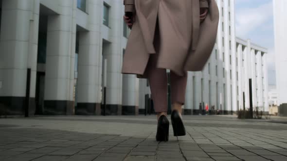 Business female legs in heels, in trousers with a developing coat, walks through the business center