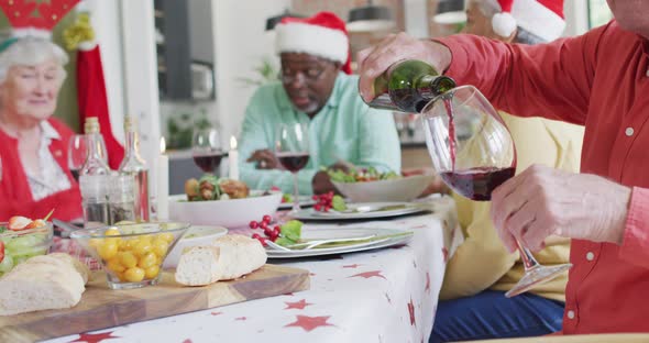 Happy diverse female and male senior friends celebrating meal, pouring vine at christmas time