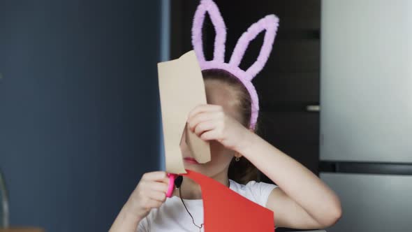 Little Girl with a Pink Bunny Ears Cuts with Scissors Objects From a Red Paper