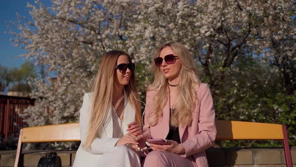 Two females persons looking at smartphone smile and laugh outside. Young adult women in sunglasses