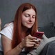 Beautiful Young 30s Redhaired Smiling Girl Lying on the Couch and Uses a Smartphone at Home Living - VideoHive Item for Sale
