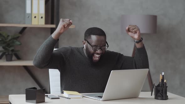 Overjoyed Black Male Student Sit at Desk Look at Laptop Screen Scream Passing Exam Getting High