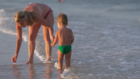 Mom Walks Along the Sea Beach with Her Son Collecting Seashells Under the Summer Sun