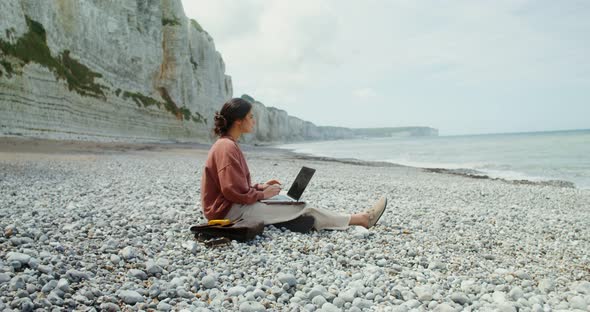 A Young Woman Uses a Laptop While Sitting on a Seashore Near Sheer Chalk Cliffs