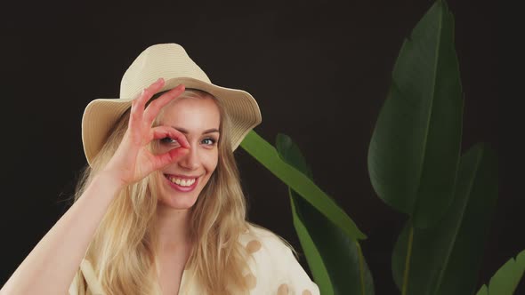 Funny European Blonde Young Girl Standing in Front of a Bigleaf Plant in a Hat Doing Ok Gesture with