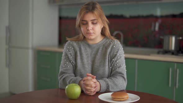 Portrait of Thoughtful Chubby Woman Alternately Looking at the Burger and Green Apple in the Kitchen