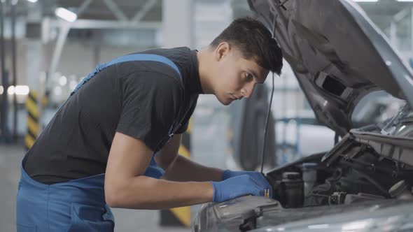 Side View of Young Brunette Caucasian Auto Mechanic Looking at Car Engine in Open Hood and Thinking