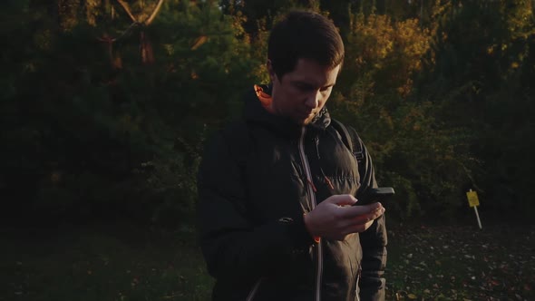 Young Man in Black Jacket in the Autumn Park Surf the Internet Using Smartphone
