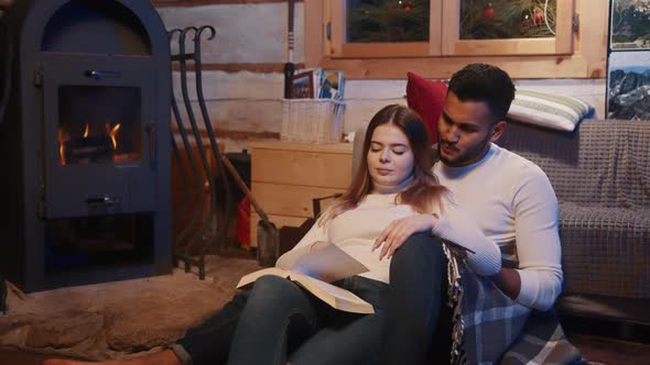 Happy Young Couple Relaxing and Reading Book Together Near the Fireplace