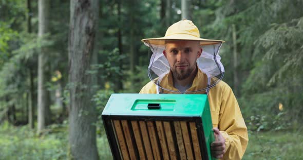 A Young Experienced Smiling Man Engaged in Beekeeping Holds an Apiary of Bees in His Hands Looks at