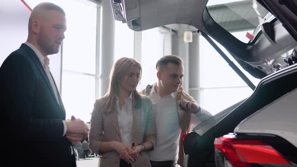 Professional Car Salesman is Telling Interested Buyers Couple About Luxurious Car with Trunk Open in