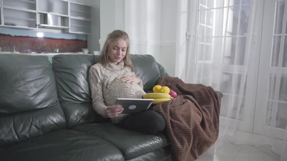 Portrait of Happy Pregnant Caucasian Woman Watching Video on Tablet and Caressing Her Belly
