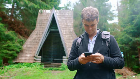 Young Man with a Backpack Stands in the Forest and Writes a Message on the Phone