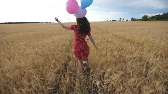 Happy Girl in Red Dress Running Through Golden Wheat Field with Balloons in Hand at Overcast Day