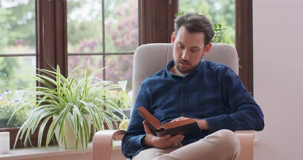 Close Up Portrait of Young Bearded Man Reading Book While Sitting in White Chair with One Leg