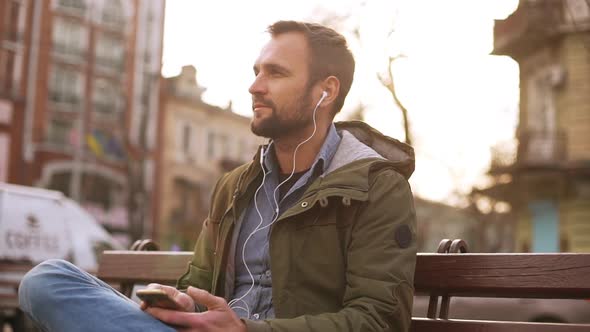 Handsome Man Listening Music in Earphones in City Park Sitting on Bench