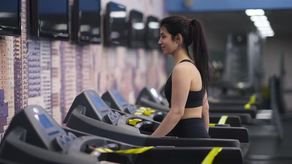 Side View of Smiling Fit Middle Eastern Young Woman Walking on Treadmill in Gym