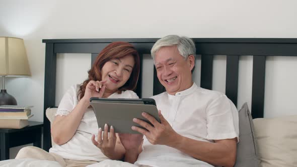 Asian senior couple using tablet after wake up, watching movie lying on bed in bedroom at home.