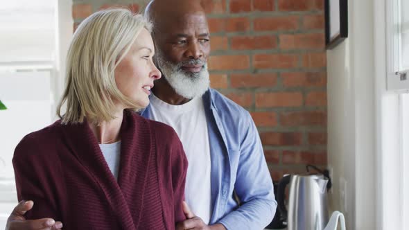 Happy senior diverse couple in kitchen, looking through window and embracing