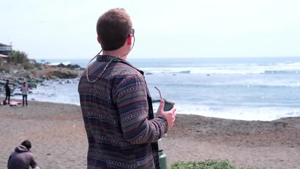 Young blond surfer man drinks Argentine mate while watching the sea and waves before surfing Argenti