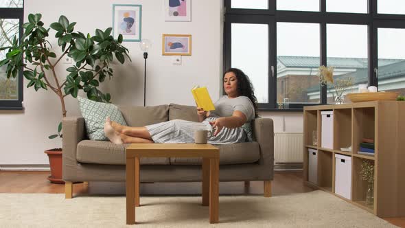 Happy Young Woman Reading Book on Sofa at Home