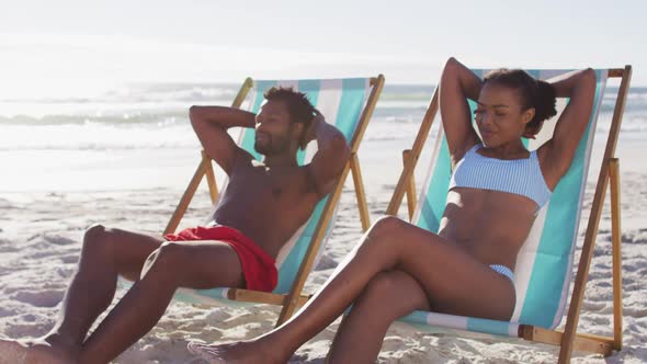 African american couple sunbathing while sitting on deck chairs at the beach