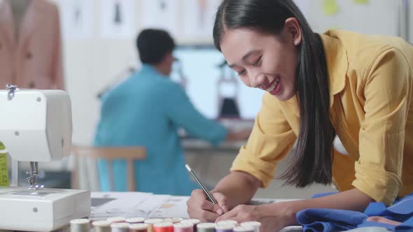 Close Up Of Asian Female Drawing Clothes On The Paper While A Male Designing Clothes On The Desktop