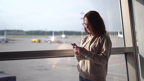 Young Woman with a Phone in Her Hands on the Background of a Window at the Airport. Airplanes in the