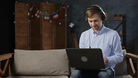 Smiling young man in headphones using laptop while sitting on sofa at home