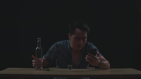 Drunk, Depressed Asian Man Drinking Vodka From Glass During Using Smartphone In Black Background